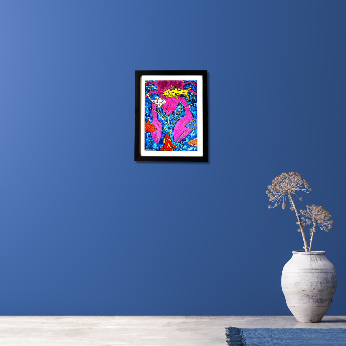 Framed colorful abstract painting on a blue wall with a vase of dried flowers below.